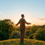 Homme debout dans une prairie au coucher du soleil, les bras ouverts vers le ciel, symbolisant la libertĂ© et le bien-ĂȘtre.